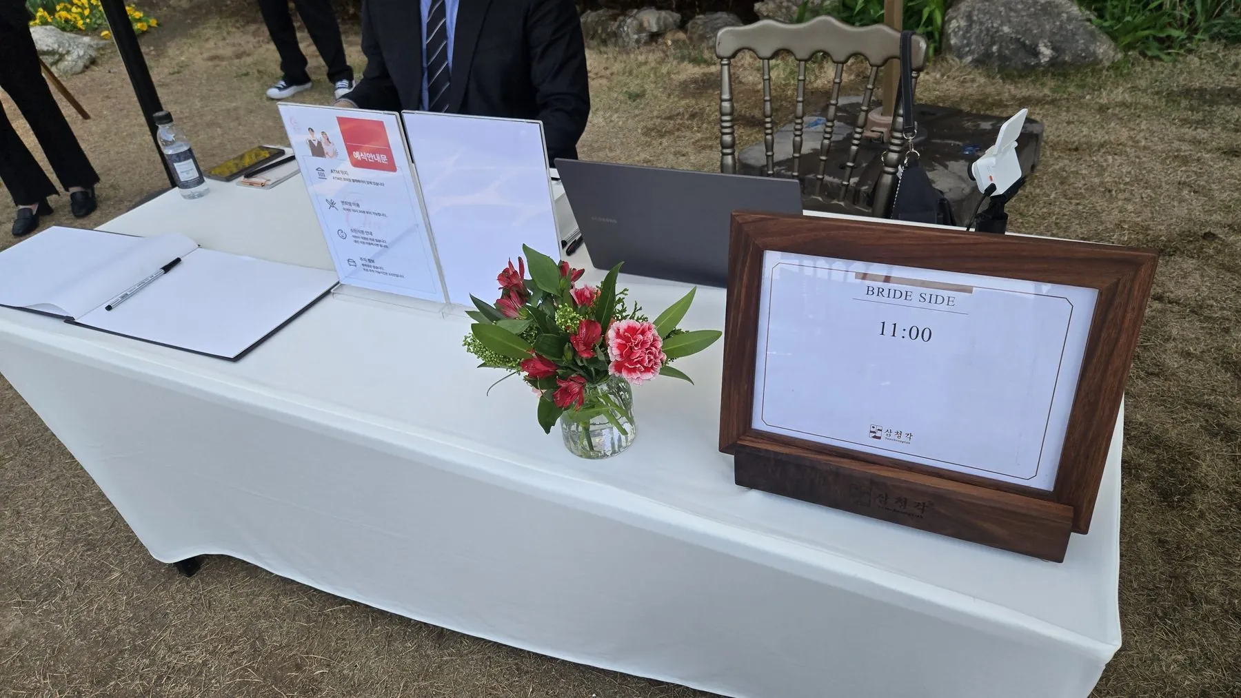 야외 가든 예식의 신부측 축의대. 흰 테이블보 위에 방명록, 예식안내문, 꽃병, 명패가 놓여 있다