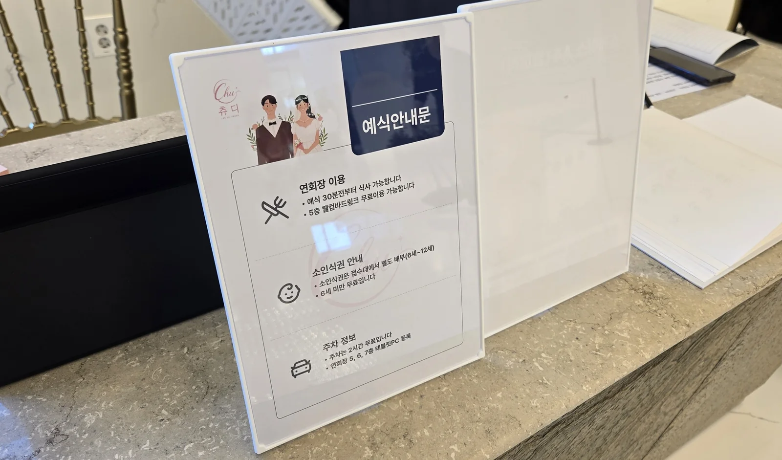 축의대 위 아크릴 거치대에 세운 예식안내문. 연회장 이용, 소인식권, 주차 정보를 안내한다